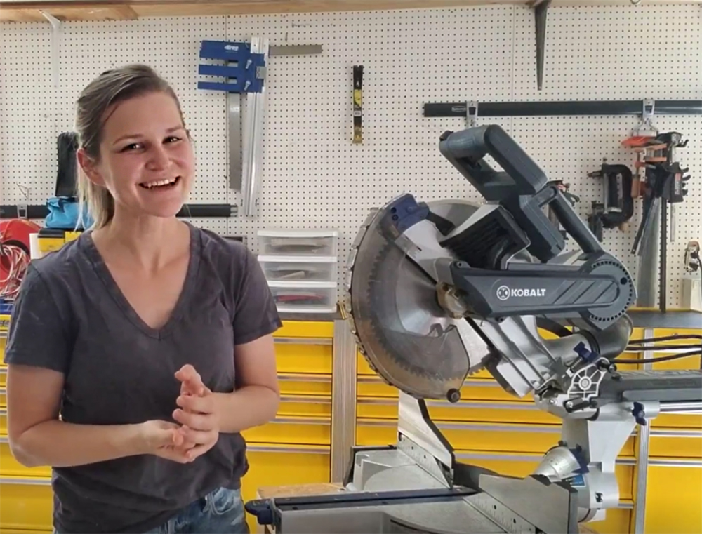 Emilee stands in a workshop with tool benches, tools and a saw