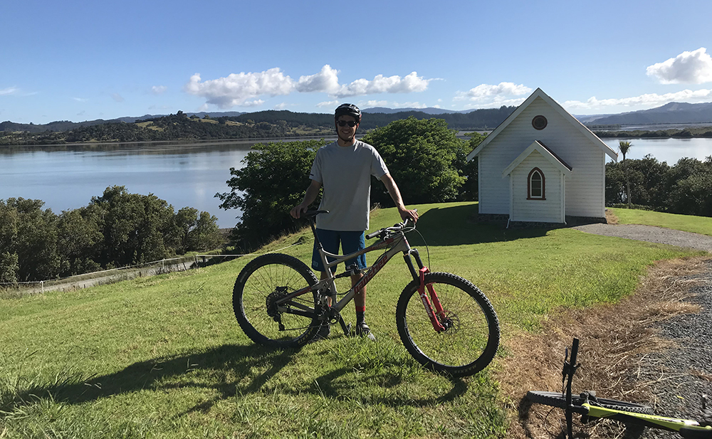 Patrick stands with his bicycle outdoors with water and landscape in the backgrounf