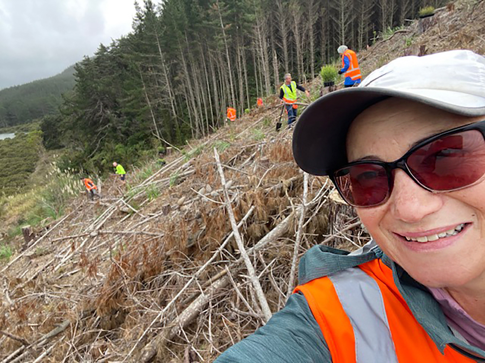 Woman takes selfie of herself with a large group of people planting trees on a steep hillside