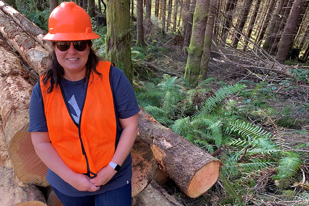 Tracie Gibbs standing in front of a pile of logs wearing orange vest and hard hat