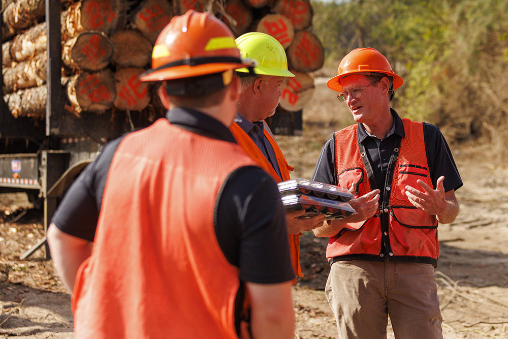 Three men in hard hats talking in front of a loaded log truck